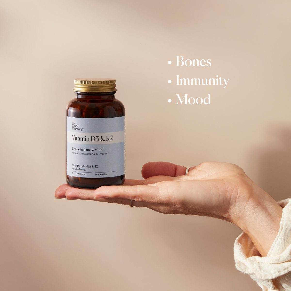 A hand holds a dark amber supplement bottle with a gold cap and a white label reading Vitamin D3 & K2. The background is a soft warm beige, with white text on the right listing benefits: Bones, Immunity, and Mood.