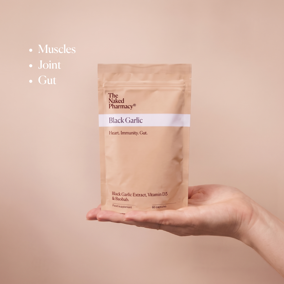 A hand holding a stand-up pouch of The Naked Pharmacy's Black Garlic supplement against a solid beige background, with bullet points listing 'Muscles', 'Joint', and 'Gut' on the left.