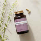 An amber supplement bottle labeled 'Immune Hero' by The Good Pharmacy, styled on a light textured surface with a gold cap, two capsules nearby, and green plant stems on the left.