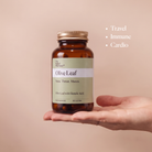 A hand holding an amber supplement bottle labeled 'Olive Leaf' by The Naked Pharmacy, set against a warm beige background with bullet points reading 'Travel', 'Immune', and 'Cardio'.