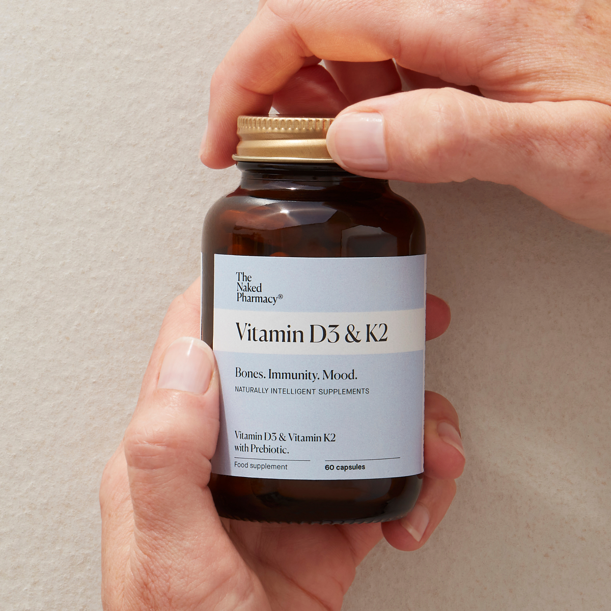 A person's hands holding a dark amber supplement bottle with a white label reading The Naked Pharmacy Vitamin D3 & K2'. The bottle has a gold screw cap and is photographed against a plain, light beige textured background.