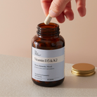 An amber glass supplement bottle labeled The Naked Pharmacy Vitamin D3 & K2'  with a hand dropping a white capsule into it, on a beige surface with a gold cap beside it.
