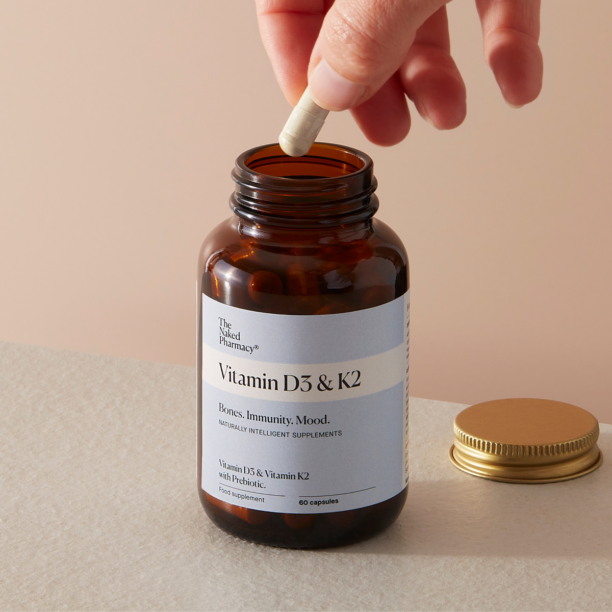 An amber glass supplement bottle labeled The Naked Pharmacy Vitamin D3 & K2'  with a hand dropping a white capsule into it, on a beige surface with a gold cap beside it.